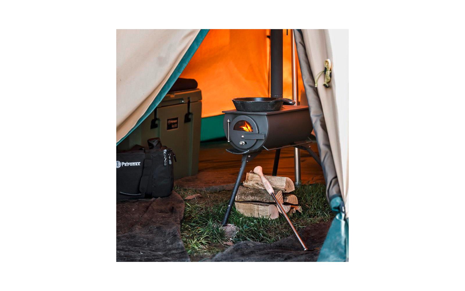 Offenes Zelt mit kleinem Metallofen, sichtbare Flamme, Pfanne auf dem Ofen, gestapelte Holzscheite mit Axt, Kühlbox und Tasche am Eingang