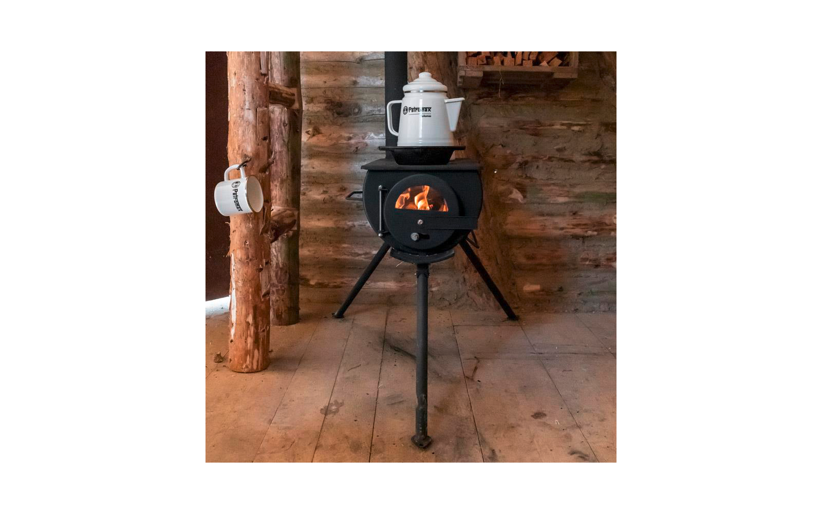 Schwarzer, runder Holzofen auf drei Beinen mit sichtbarer Flamme, weißer Kessel auf dem Ofen, weißer Becher an einem Holzbalken, Holzblockwand und Holzboden