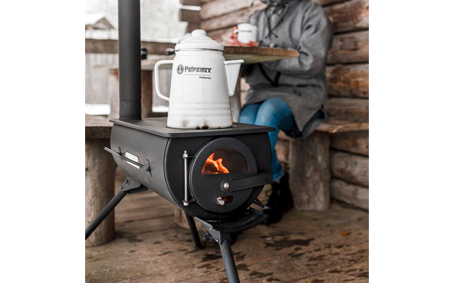 Schwarzer Holzofen mit sichtbarer Flamme, weißer Kessel 'Petromax' darauf, sitzende Person in grauem Mantel mit Tasse auf Holzbank, Wände aus Rundholz