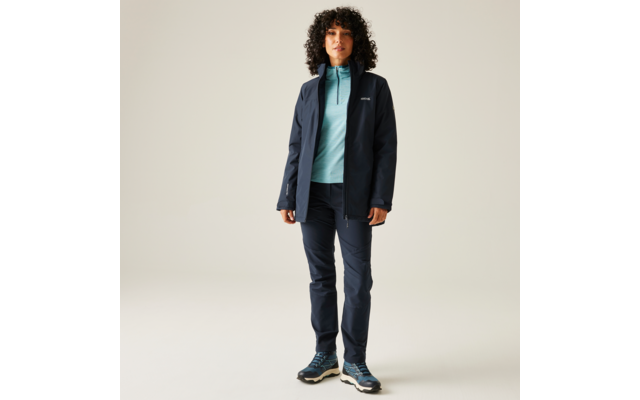Homme aux cheveux bouclés portant un jean bleu, un pull bleu clair et un manteau sombre, debout devant un fond clair