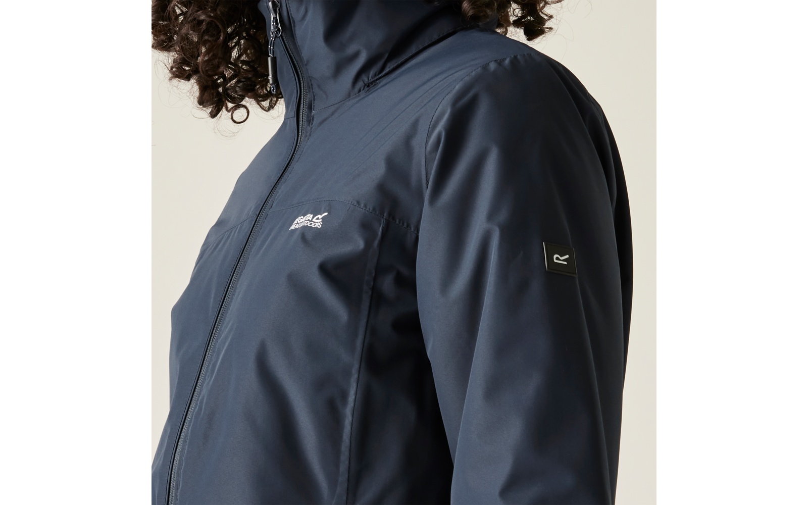 Dunkelblaue Jacke mit Stehkragen und durchgehendem Reißverschluss, kleines Logo auf linker Brust, kleines quadratisches Label am rechten Oberarm, lockige Haare am oberen Bildrand, heller Hintergrund