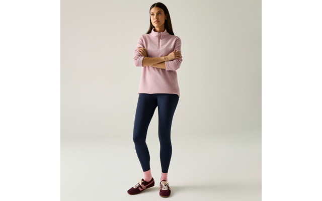 Persona in piedi con le braccia incrociate, indossa un maglione rosa con mezza zip, leggings blu scuro, calzini rosa e sneaker bordeaux su sfondo chiaro e uniforme