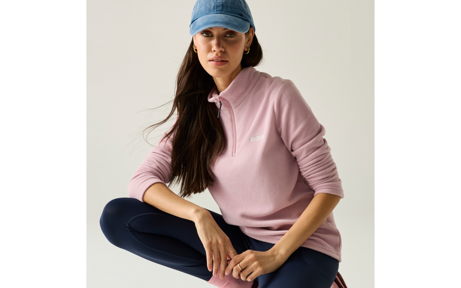 Persona seduta con un ginocchio sollevato su sfondo chiaro, indossa una felpa rosa con zip corta, leggings blu scuro, un berretto da baseball blu e lunghi capelli scuri