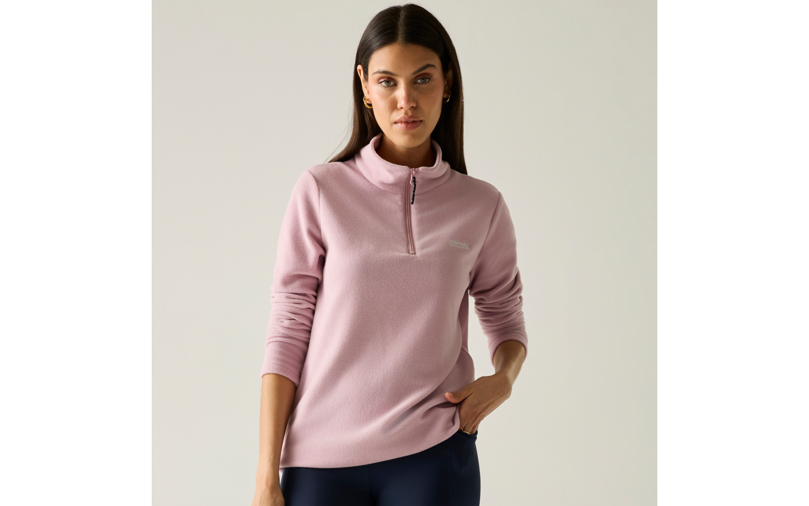 Persona con capelli lunghi e scuri che indossa una maglia rosa con mezza cerniera, una mano in tasca, sfondo chiaro