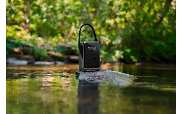Schwarze tragbare Powerstation mit digitalem Display und Tragegriff auf einem Felsen in einem flachen Bach, grüne Bäume im Hintergrund