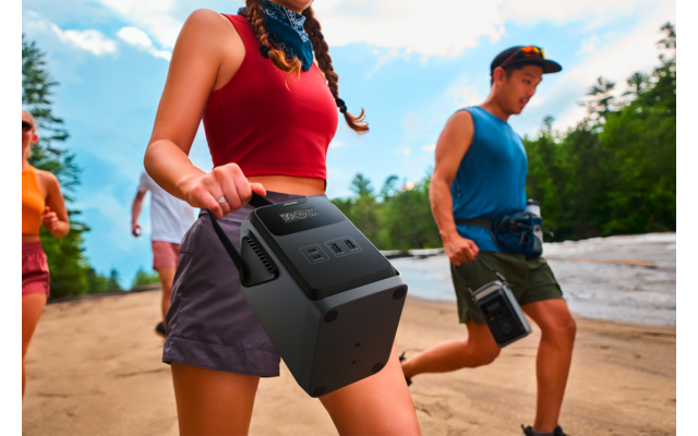 Mehrere Personen auf sandigem Ufer neben Wasser; im Vordergrund eine Frau in rotem Top mit geflochtenem Zopf hält ein schwarzes tragbares Gerät am Griff; im Hintergrund zwei Personen, Bäume und Himmel