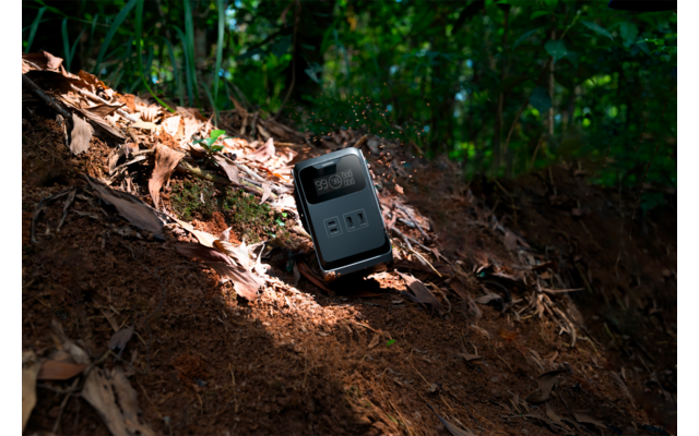 Schwarzes rechteckiges elektronisches Gerät mit digitaler Anzeige '100%' und zwei Anschlüssen, auf schrägem Erd- und Laubboden im Wald, teils von Sonnenlicht beleuchtet