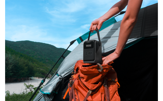 Hände platzieren ein kleines schwarzes tragbares Ladegerät mit Display auf einem orangefarbenen Rucksack am Eingang eines Zeltes; See, bewaldete Hügel und blauer Himmel im Hintergrund
