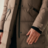 Eine Person trägt eine beige Winterjacke und legt die rechte Hand auf den Körper