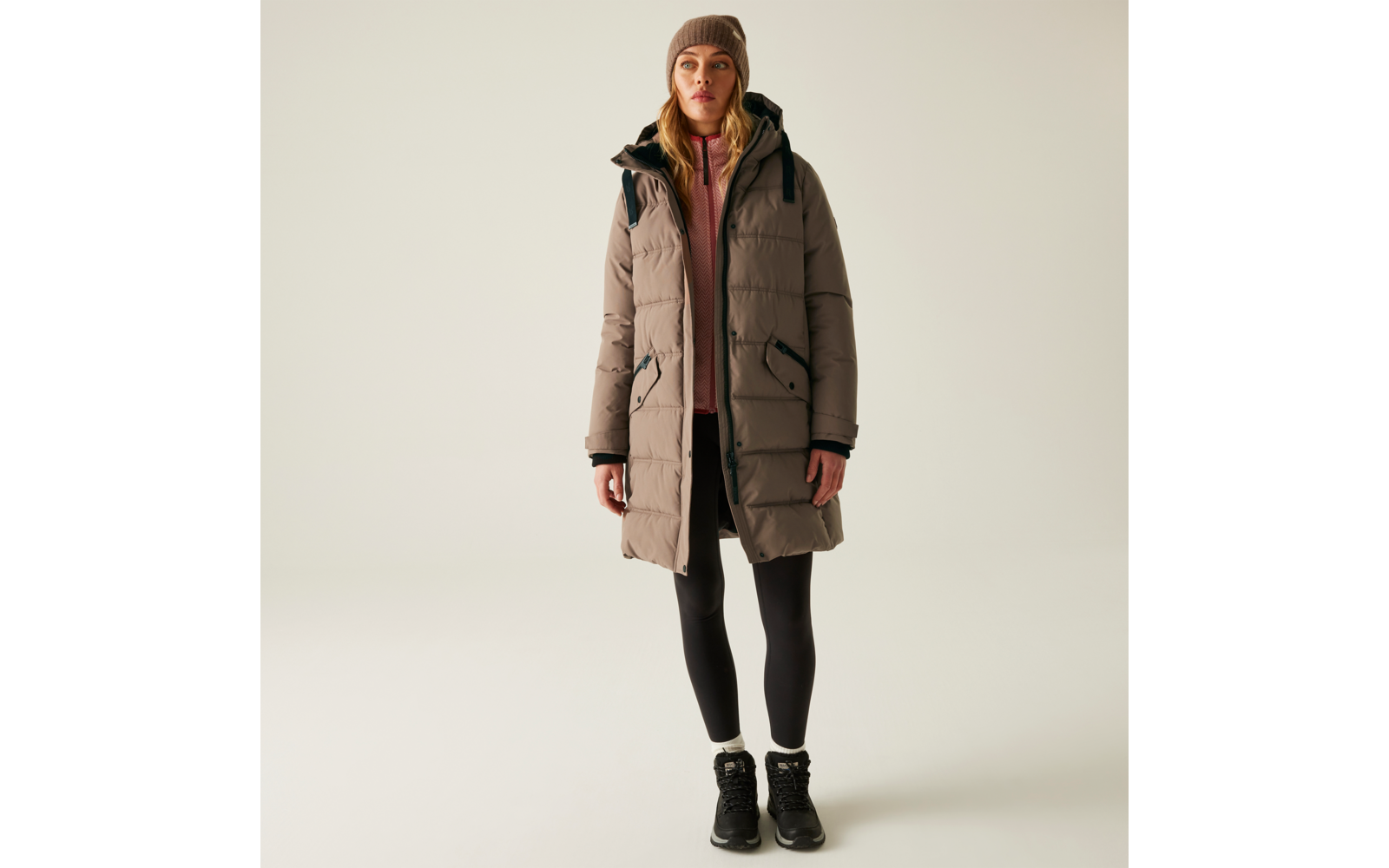Frau trägt lange winterliche Jacke mit Kapuze, darunter schwarze Hose und Schuhe, vor hellem Hintergrund