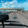 Blick aus einem Fahrzeug auf mehrere Fahrspuren und Zahlstellen unter einer Überdachung, einige Autos, blauer Himmel mit Wolken, Armaturenbrett und Scheibenwischer im Vordergrund