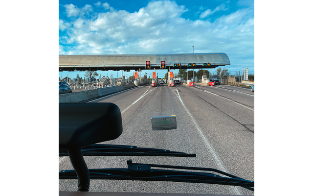 Blick aus einem Fahrzeug auf mehrere Fahrspuren und Zahlstellen unter einer Überdachung, einige Autos, blauer Himmel mit Wolken, Armaturenbrett und Scheibenwischer im Vordergrund