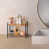Zweistöckiges Holzregal mit schwarzen Metallstützen vor beiger Wand neben der Kante einer Badewanne und einem runden Spiegel; oben drei weiße/goldene Flaschen, ein tropfenförmiges Glasfläschchen und ein rosa Behälter mit Holzdeckel; unten eine dunkle