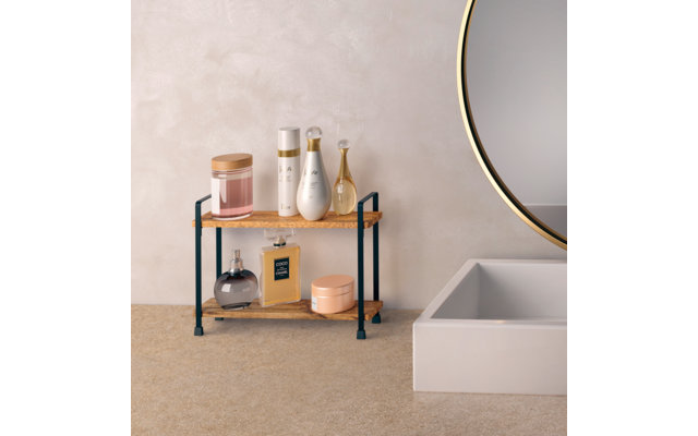 Zweistöckiges Holzregal mit schwarzen Metallstützen vor beiger Wand neben der Kante einer Badewanne und einem runden Spiegel; oben drei weiße/goldene Flaschen, ein tropfenförmiges Glasfläschchen und ein rosa Behälter mit Holzdeckel; unten eine dunkle