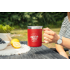 Main tenant une tasse isotherme rouge avec logo blanc 'camp life' sur une table en bois foncé ; à côté un bol de myrtilles, un demi-citron et un citron entier, un petit pot avec une plante ; arrière-plan vert flou ; main avec manche rayée et montre-bracelet