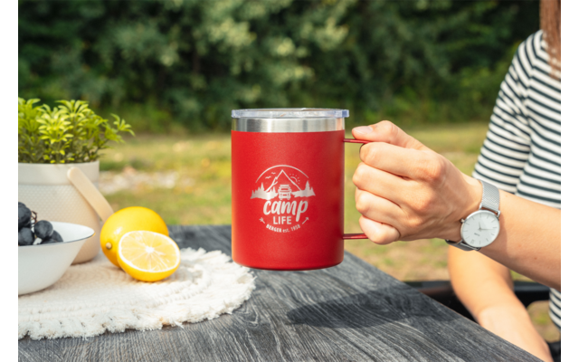 Main tenant une tasse isotherme rouge avec logo blanc 'camp life' sur une table en bois foncé ; à côté un bol de myrtilles, un demi-citron et un citron entier, un petit pot avec une plante ; arrière-plan vert flou ; main avec manche rayée et montre-bracelet