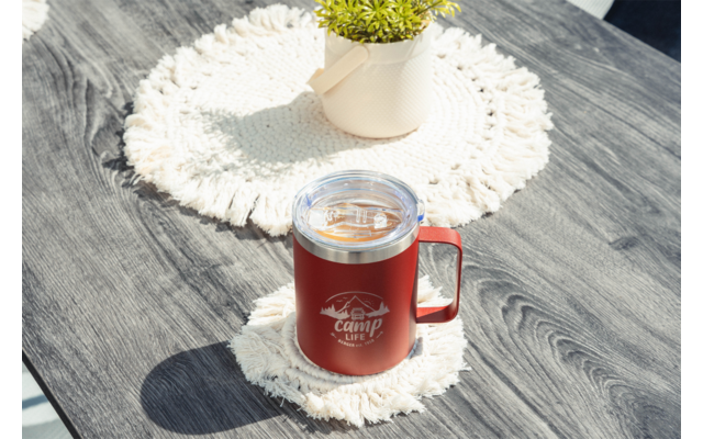 Tasse rouge avec couvercle transparent et anse, logo blanc à l'avant, posée sur un dessous de tasse crocheté blanc sur une table en bois foncé; plante en pot sur un autre dessous en arrière-plan
