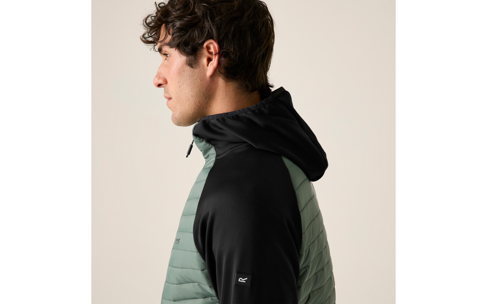 Seitenansicht einer Person mit dunklem lockigem Haar, die eine gesteppte hellgrüne Jacke mit schwarzer Kapuze und schwarzen Ärmeln trägt vor hellem Hintergrund