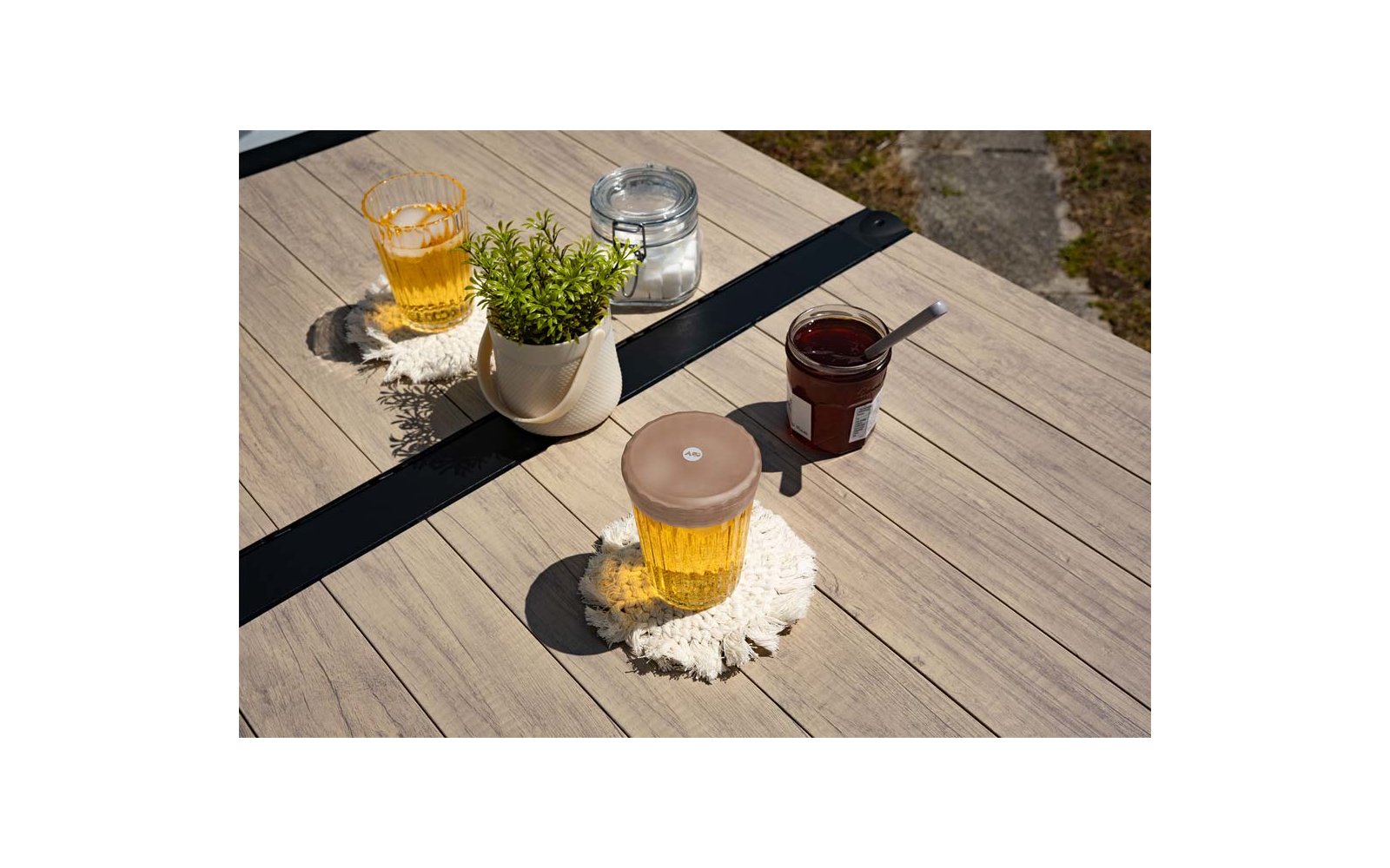 Houten tafel met twee glazen gele vloeistof op witte gehaakte onderzetters, witte plantenpot met groene plant, klein potje met donkere inhoud en lepel, klein glazen potje met beugel; stenen ondergrond op de achtergrond