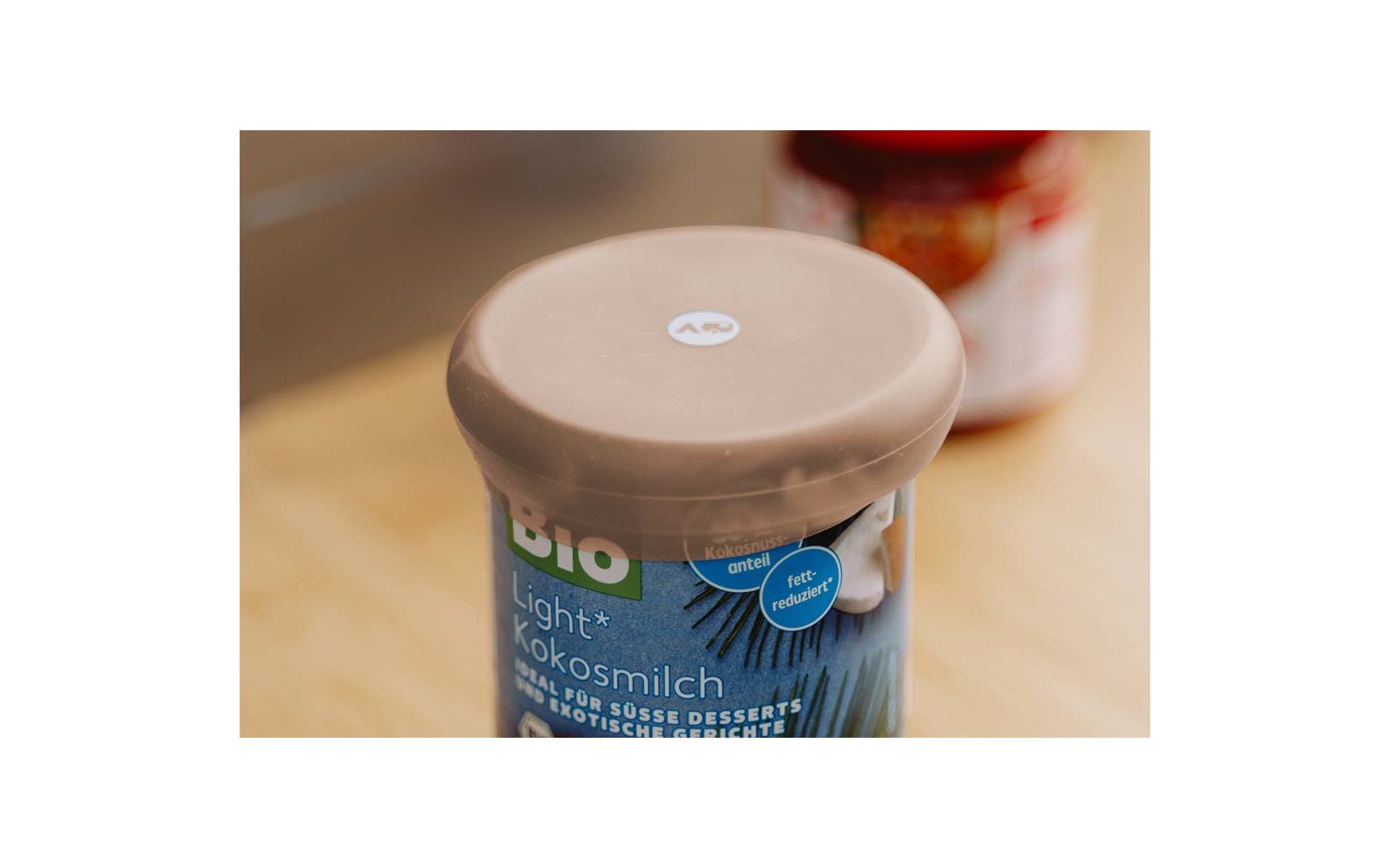 Bekertje met beige kunststofdeksel en blauw etiket met opschrift 'BIO Light Kokosmilch' op een houten ondergrond; op de achtergrond onscherpe pot met rood deksel