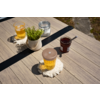 Tavolo di legno con due bicchieri di liquido giallo su sottobicchieri bianchi all'uncinetto, vaso bianco con pianta verde, piccolo barattolo con contenuto scuro e cucchiaio, piccolo barattolo con chiusura metallica; pavimento in pietra sullo sfondo