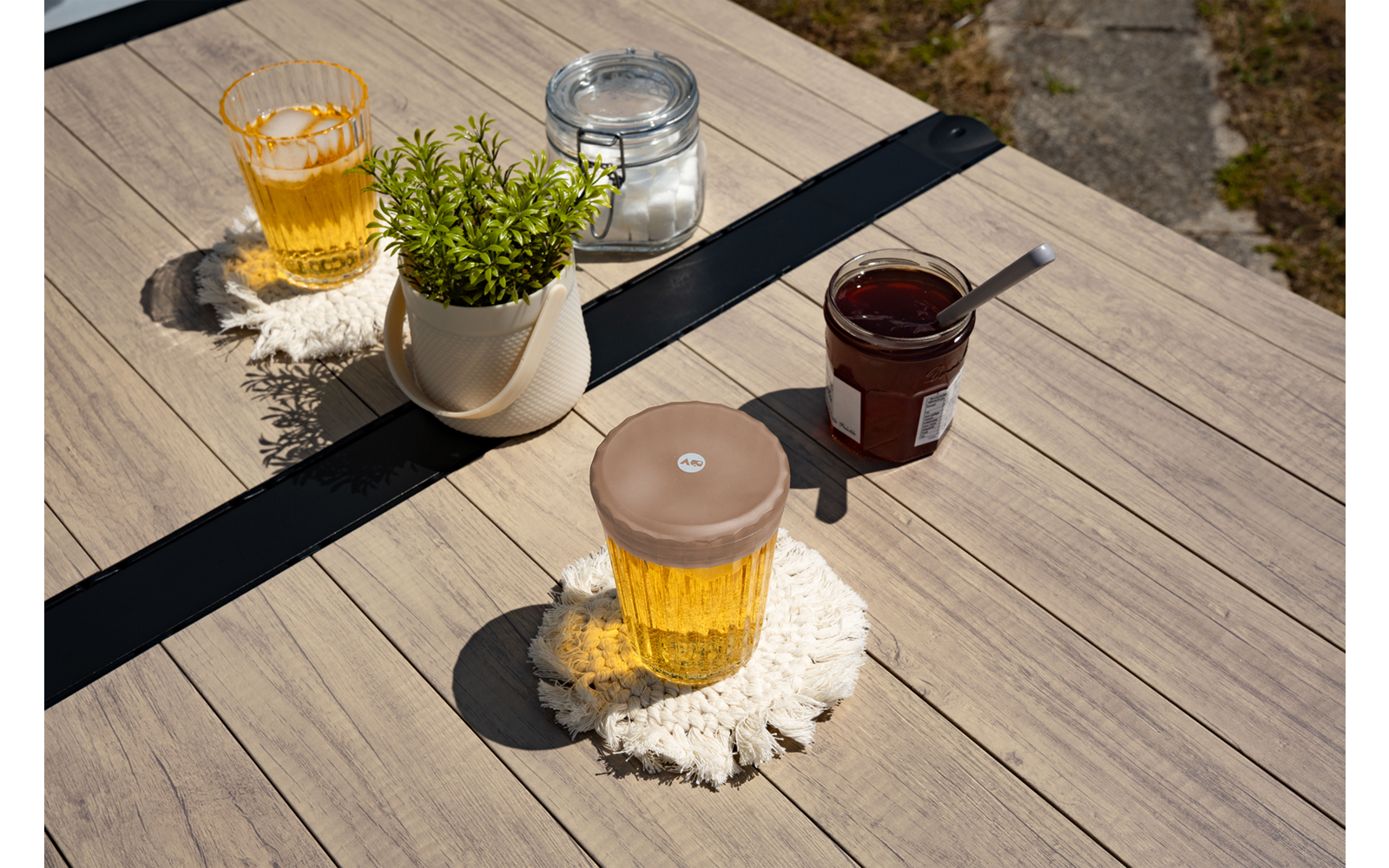 Tavolo di legno con due bicchieri di liquido giallo su sottobicchieri bianchi all'uncinetto, vaso bianco con pianta verde, piccolo barattolo con contenuto scuro e cucchiaio, piccolo barattolo con chiusura metallica; pavimento in pietra sullo sfondo