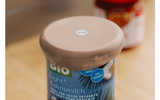 Contenitore con coperchio in plastica beige ed etichetta blu con la scritta 'BIO Light Kokosmilch' su una superficie di legno; sullo sfondo un barattolo sfocato con coperchio rosso