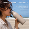 Frau mit Sonnenbrille trinkt aus beige Trinkflasche am Strand; im Hintergrund Sand, Meer und blauer Himmel; oben weißer Text 'UNBESCHWERTER GENUSS' und kleiner deutscher Text