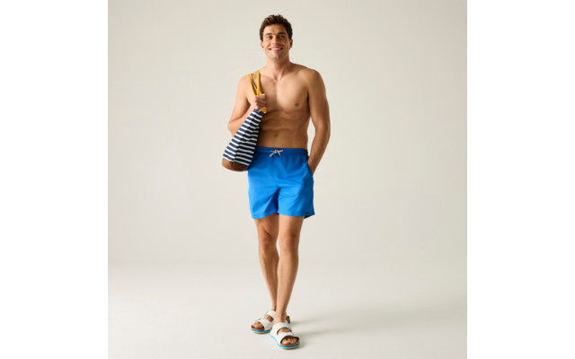 Homme torse nu en short de bain bleu et sandales blanches, porte un sac rayé bleu et blanc sur une épaule et a une main dans la poche, debout devant un fond uni clair