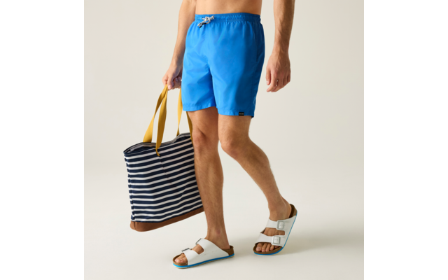 Bas du corps d'une personne debout portant un short bleu, tenant dans une main un sac rayé aux anses jaunes et portant des sandales blanches à boucles sur fond clair