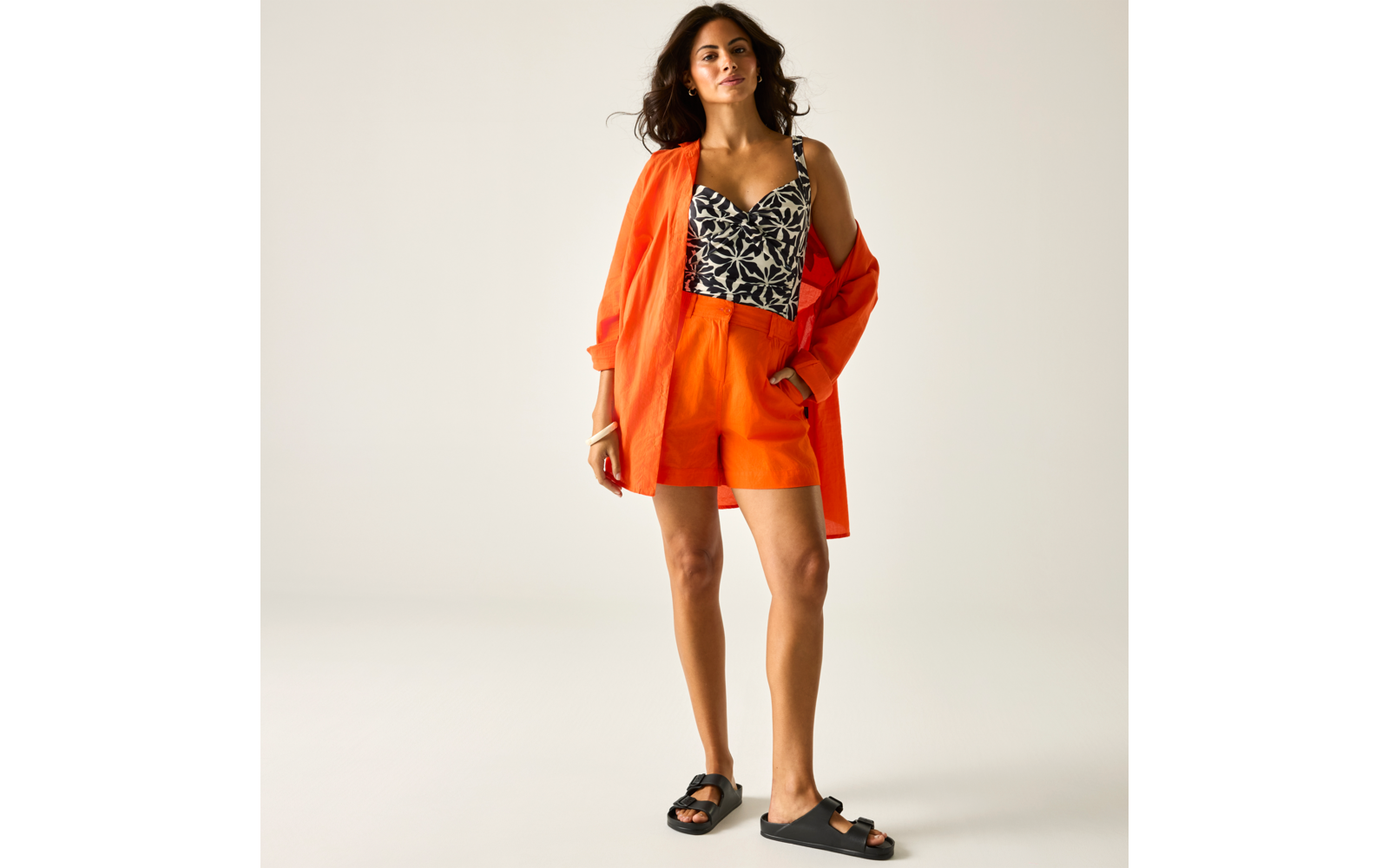 Personne debout sur un fond neutre clair, portant une veste orange ouverte et un short assorti, un top noir à motifs et des sandales noires