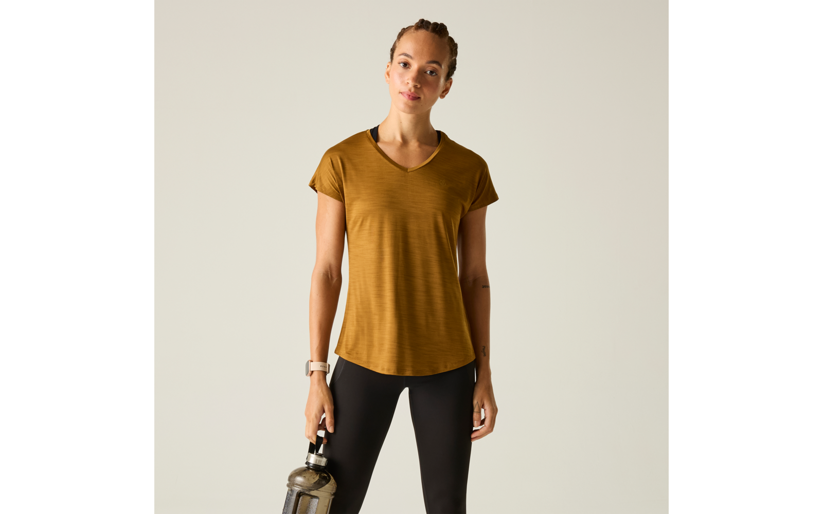 Person steht vor hellem Hintergrund, Haare zurückgebunden, trägt kurzes senffarbenes V‑Ausschnittshirt und schwarze Leggings, hält in der rechten Hand eine Trinkflasche