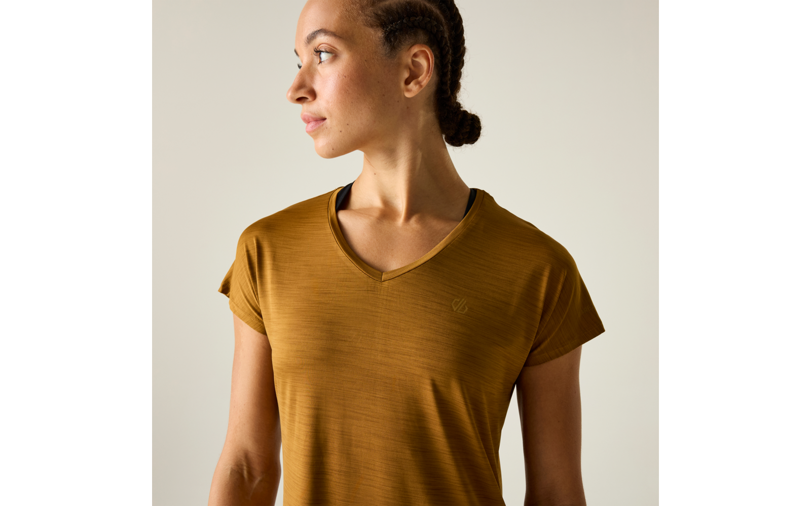 Person blickt nach links, geflochtene Haare zu einem Dutt zusammengebunden, ockerfarbenes V‑T-Shirt, einfarbiger heller Hintergrund