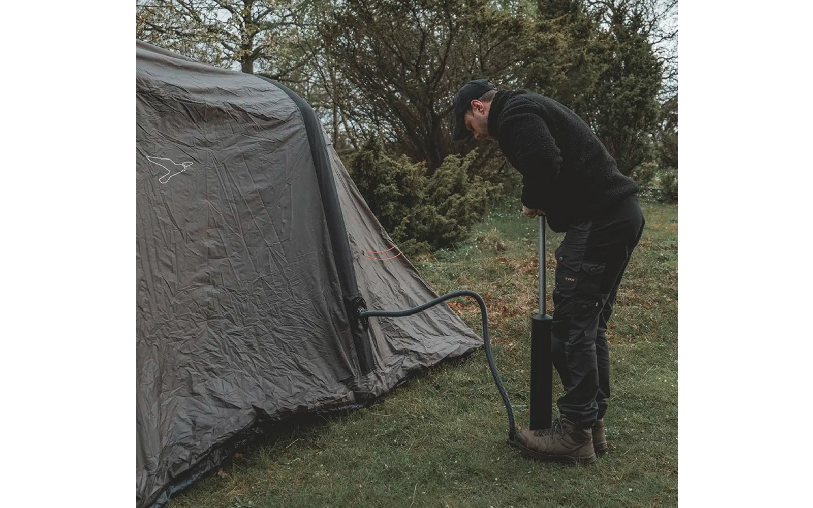 Person pumpt mit einer Luftpumpe ein graues Zelt auf, umgeben von Gras und Bäumen