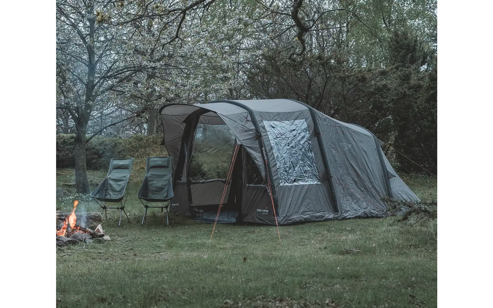 Zelt auf einer Wiese mit zwei Campingstühlen und einem Lagerfeuer daneben, im Hintergrund Bäume mit kahlen Ästen