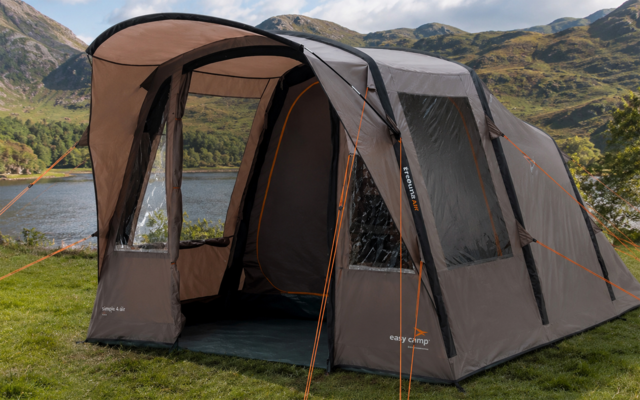 Gray camping tent with awning, clear windows and orange guy lines on grass by a lake with mountains in the background