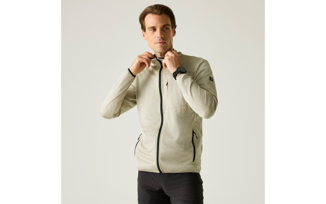 Mann in hellbeiger Jacke mit Reißverschluss zieht am Reißverschluss, steht vor hellem Hintergrund, trägt schwarze Hose und Uhr am linken Handgelenk