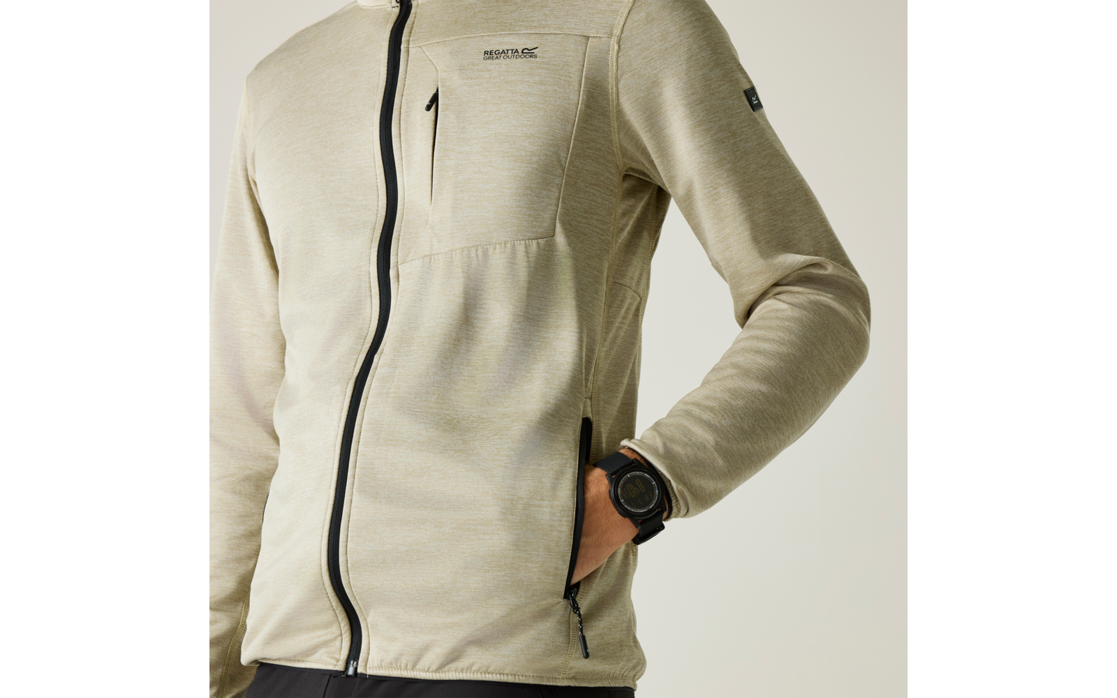 Oberkörper einer Person in hellbeiger, durchgehend gezippten Jacke mit Brusttasche, Hand in seitlicher Reißverschlusstasche und schwarzer Armbanduhr