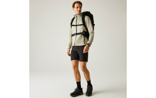 Person mit kurzem braunem Haar, hellbeiger Zippjacke, schwarzem Rucksack, schwarzen Shorts, schwarzen Wanderschuhen und Armbanduhr, steht auf hellem Hintergrund und schaut leicht nach rechts