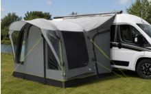Witte camperbus met grijs voortent bevestigd aan de zijkant; voortent met ramen, opgespannen en met scheerlijnen vastgezet op gras naast een vijver, bomen en blauwe lucht op de achtergrond