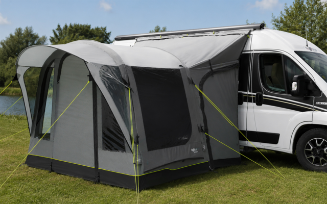 Witte camperbus met grijs voortent bevestigd aan de zijkant; voortent met ramen, opgespannen en met scheerlijnen vastgezet op gras naast een vijver, bomen en blauwe lucht op de achtergrond