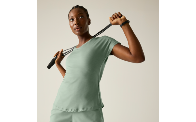 Frau vor hellem einfarbigem Hintergrund, hält ein Springseil hinter dem Nacken, trägt hellgrünes T‑Shirt und Shorts, geflochtene Haare, Smartwatch am Handgelenk, blickt nach oben rechts