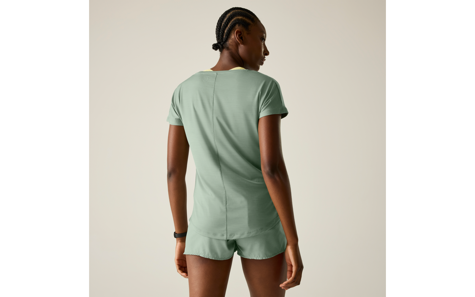 Person von hinten, trägt hellgrünes T‑Shirt und passende kurze Shorts, Haare in Cornrows geflochten und zu kleinem Dutt gebunden, steht vor einfarbig hellem Hintergrund, schwarzes Armband am linken Handgelenk