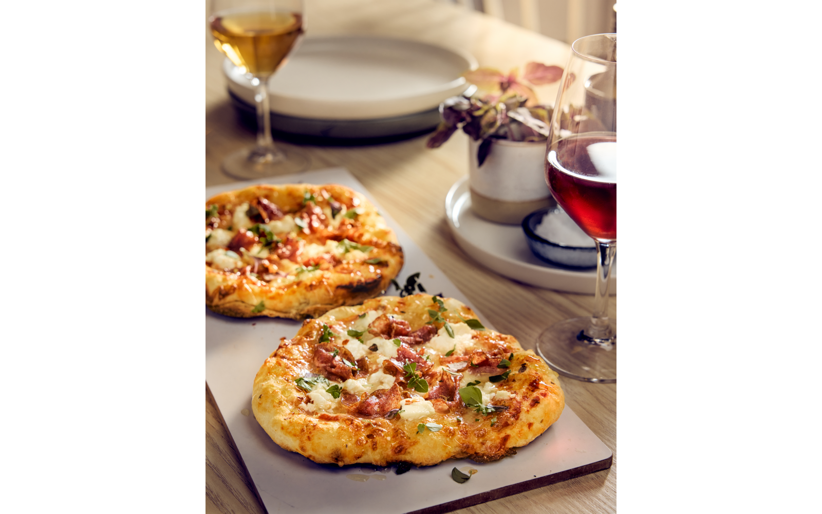 Zwei kleine Pizzen auf einem Brett, Gläser mit Rot- und Weißwein, gestapelte Teller, Topfpflanze auf Holztisch