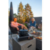 Mann in rosa Hemd und Frau in blauem Oberteil sitzen auf Stühlen auf einer Holzterrasse; im Vordergrund ein tragbarer Gaskocher mit Topf auf einem Betonblock, rechts eine Feuerstelle mit Flammen und ein Teller mit belegten Brotscheiben; im Hintergrun