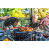 Zwei Hände heben Deckel und Pfannenwender mit einer Pizza über einem Tischgrill; im Hintergrund unscharfe Person mit blondem Haar, auf dem Tisch Gläser und Teller mit Pilzen und Gemüse