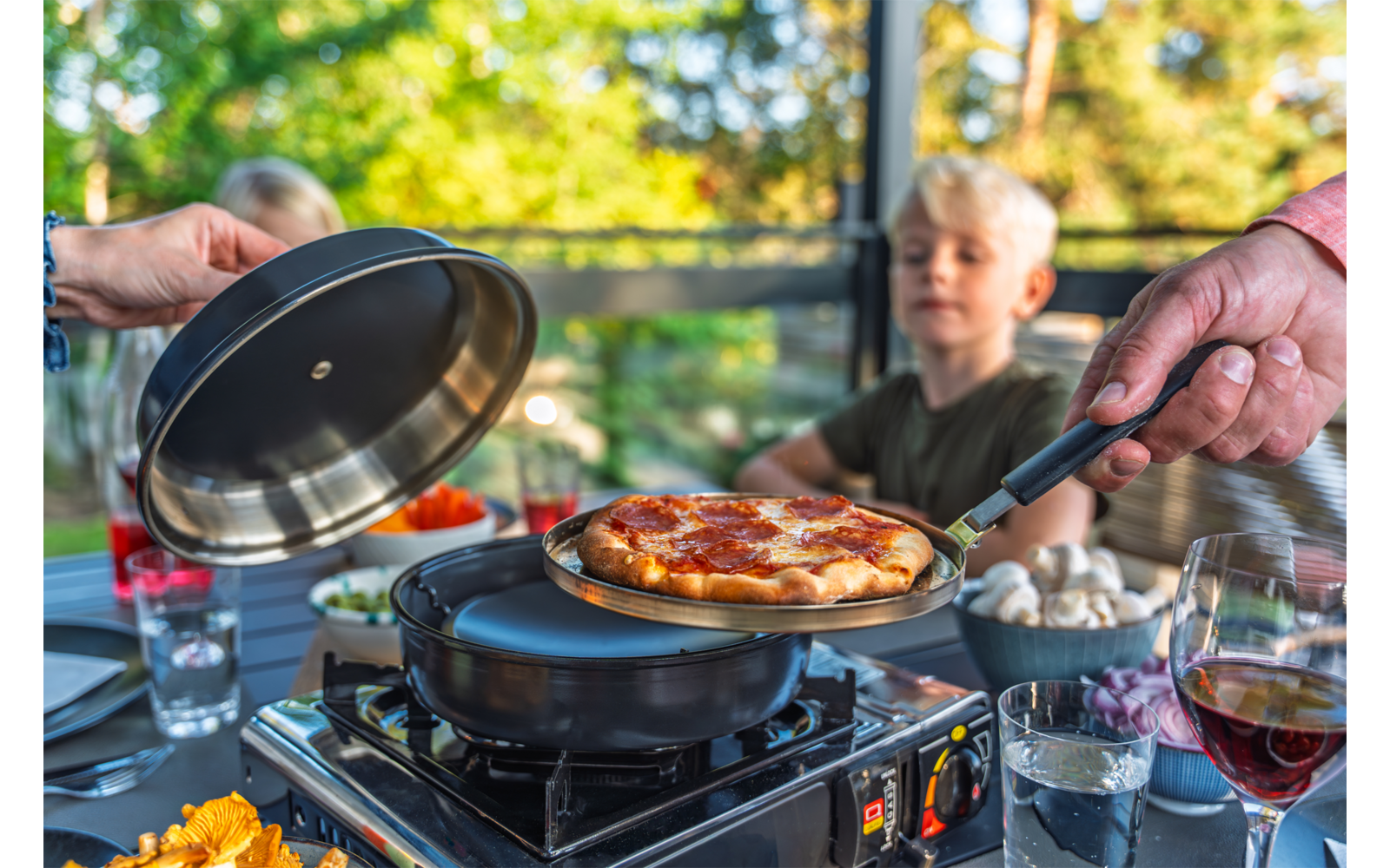 Zwei Hände heben Deckel und Pfannenwender mit einer Pizza über einem Tischgrill; im Hintergrund unscharfe Person mit blondem Haar, auf dem Tisch Gläser und Teller mit Pilzen und Gemüse