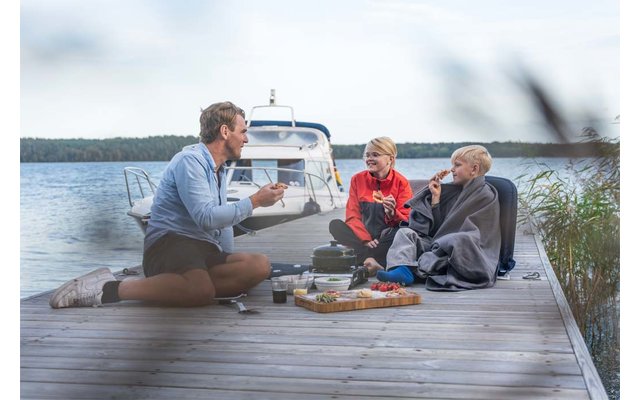 Drei Personen sitzen auf einem Holzsteg am See neben einem weißen Motorboot; auf dem Steg liegen Decke, Tassen und ein Tablett mit Snacks; eine Person ist in eine Decke gewickelt