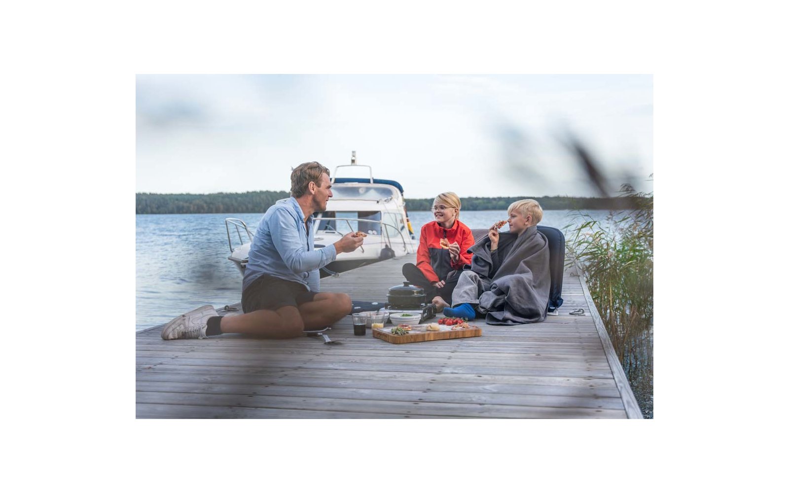 Drei Personen sitzen auf einem Holzsteg am See neben einem weißen Motorboot; auf dem Steg liegen Decke, Tassen und ein Tablett mit Snacks; eine Person ist in eine Decke gewickelt