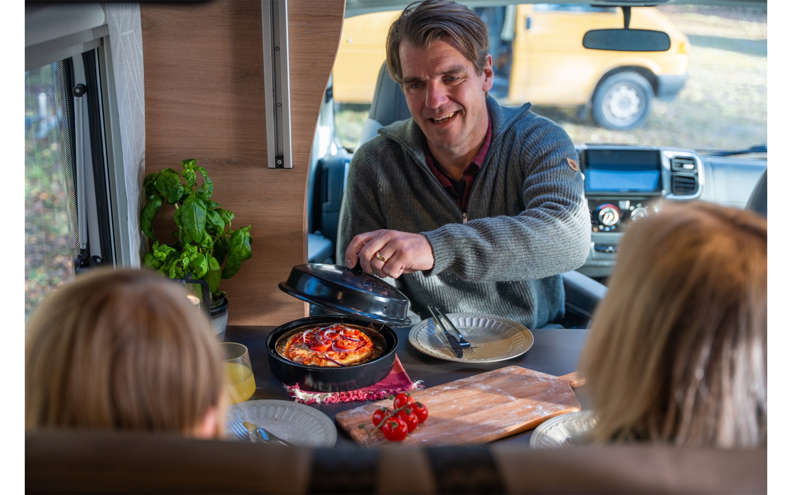 Mann in einem Wohnmobil hebt den Deckel einer Pfanne mit Pizza; zwei Personen sitzen am Tisch mit dem Rücken zur Kamera; auf dem Tisch Teller, Kirschtomaten, ein Schneidebrett und ein Basilikumtopf; durch die Windschutzscheibe ist ein geparktes Auto 